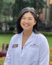 Minda Liu from the waist up, who has short dark hair, is wearing a white medical coat, and is smiling widely at the camera. Minda is standing outside with trees and grass blurred in the background.