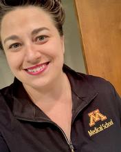 Katie McLaughlin, a white woman with brown hair pulled up and wearing a black quarter-zip with the UMN logo, looks at the camera, smiling widely. 