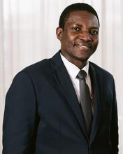 Adebayo Ibikunle, a Black man with short dark hair wearing a suit and tie, looks at the camera and smiles