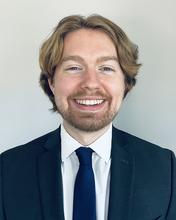 Smiling, young adult, caucasian male with medium-length strawberry-blonde hair parted just off center, and stubbly facial hair wearing a white dress shirt with blue necktie and a blue suit jacket.